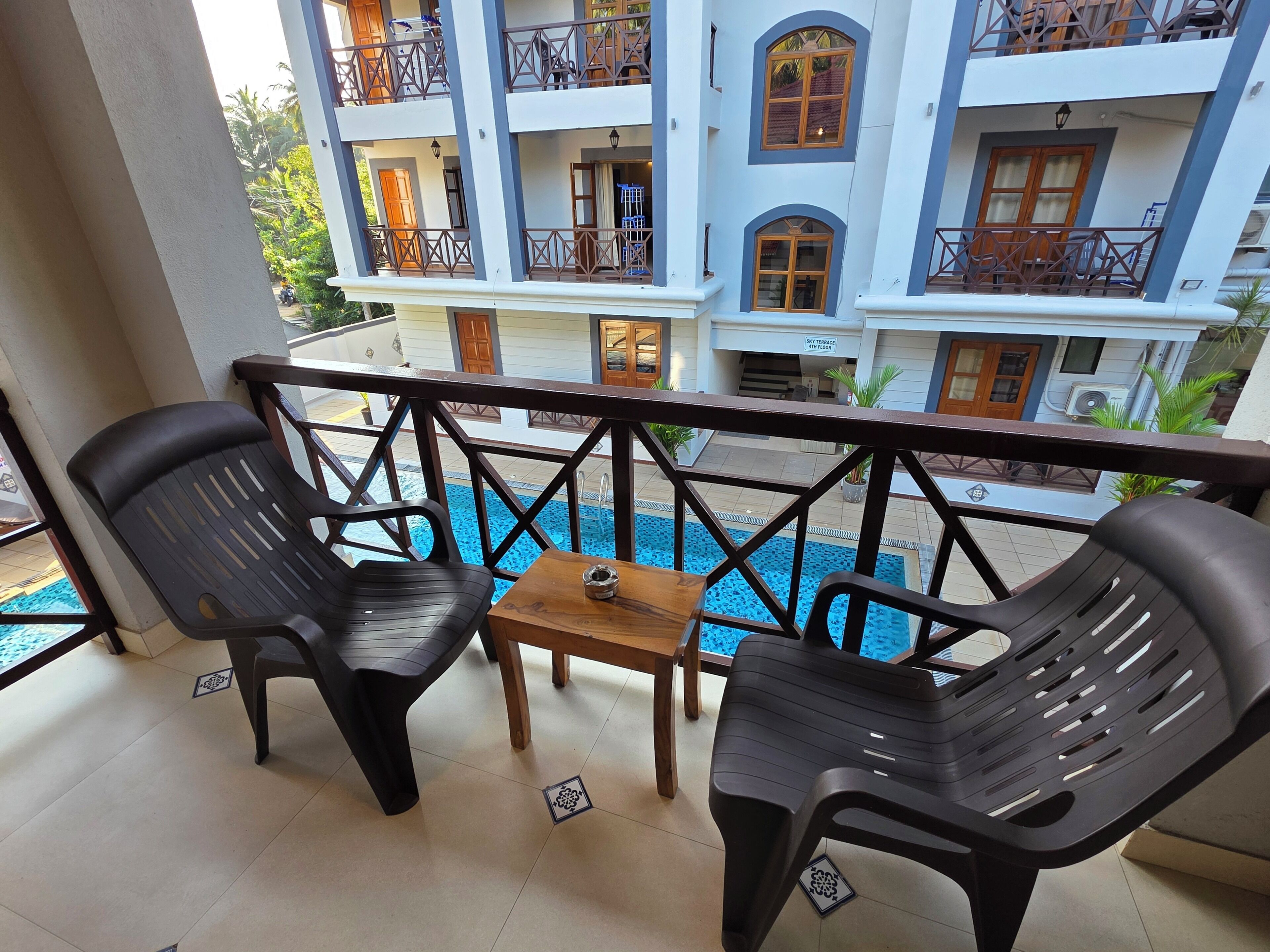 Een balkon met twee zwarte plastic stoelen, een kleine houten tafel en uitzicht op een zwembad en een meerlaags gebouw met balkons.