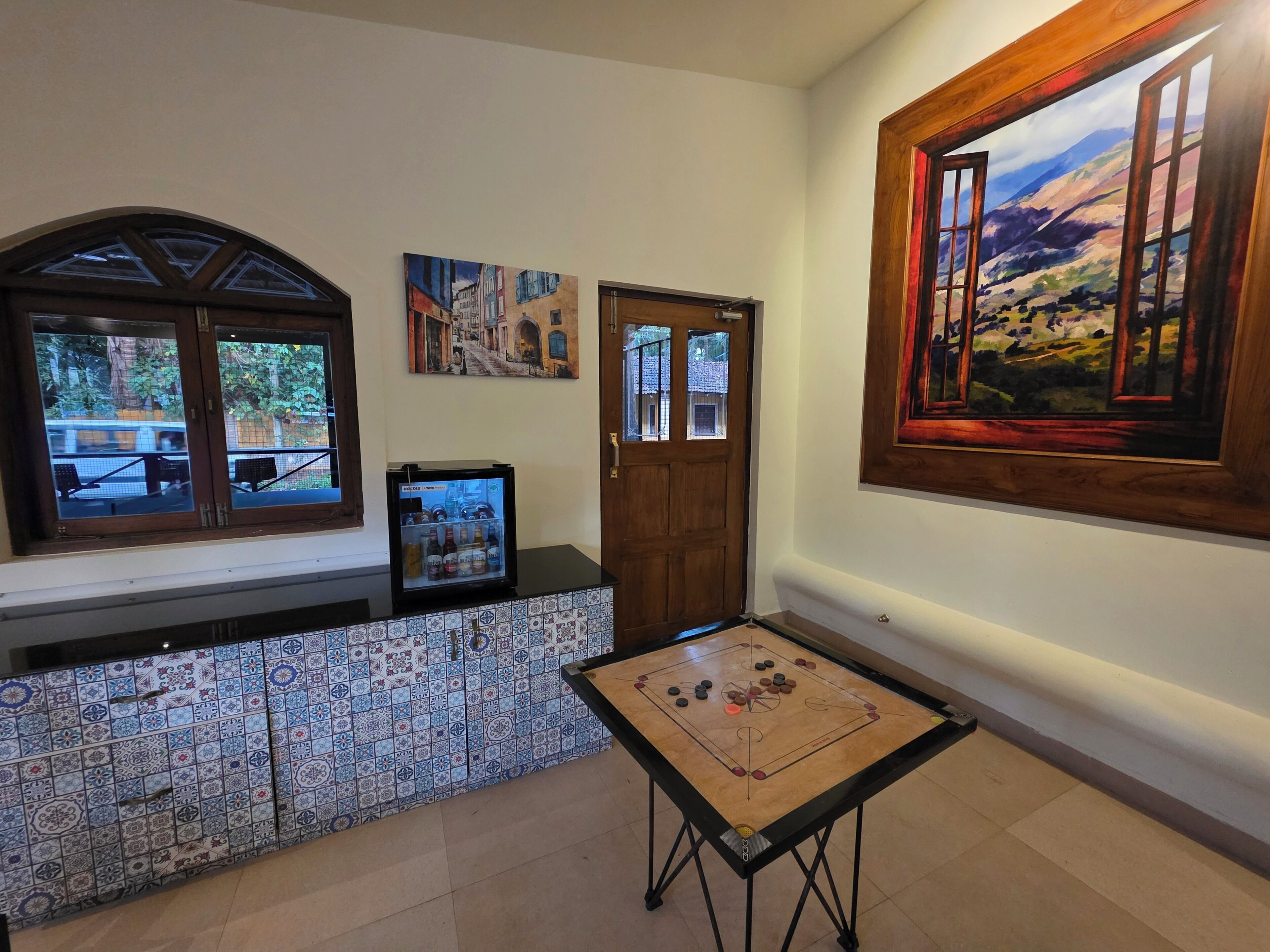 A room with a tiled wall, a door, a window, a small table with a board game, and two paintings.