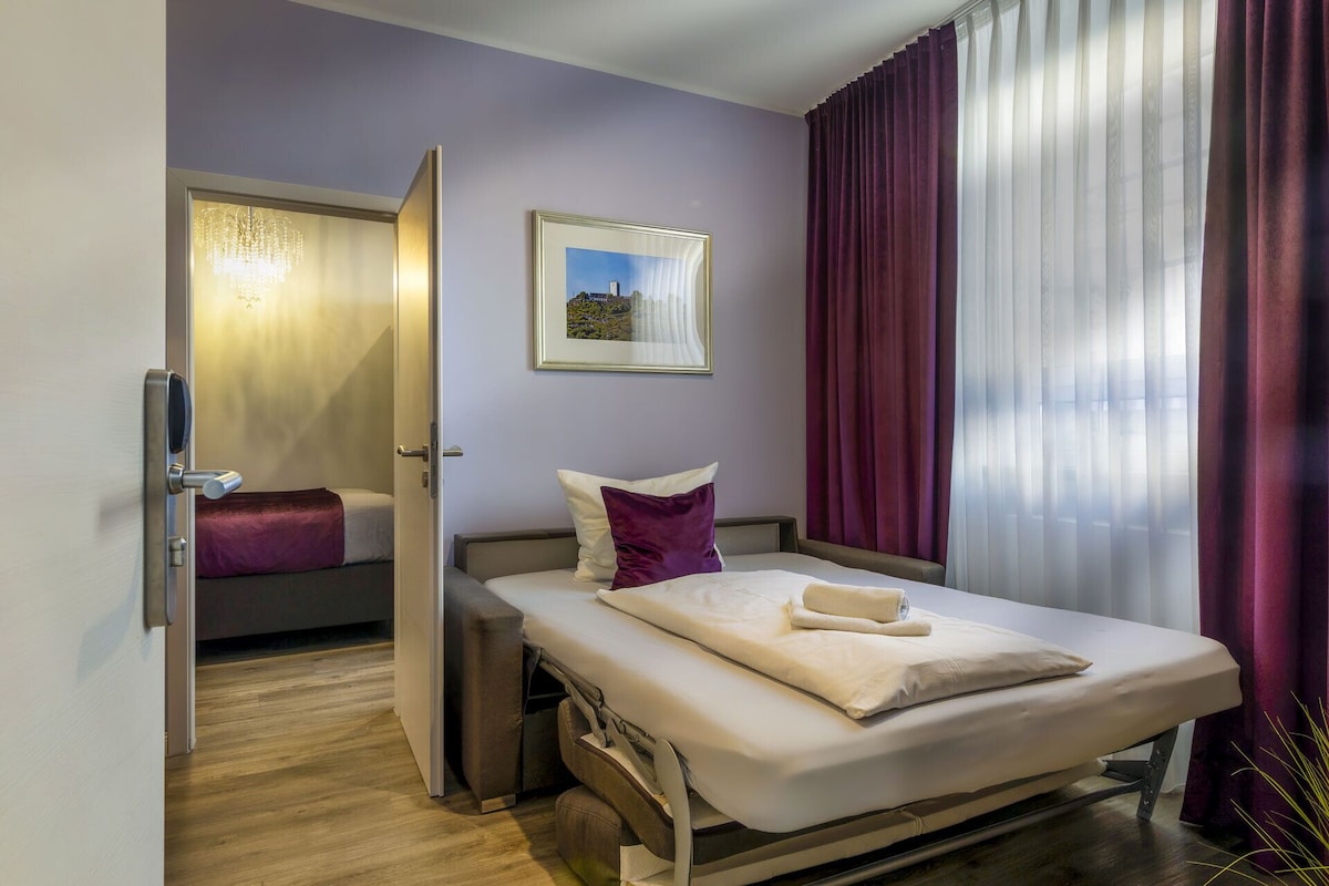 A hotel room with a bed, a bedside table, a chair, a window with curtains, and a painting on the wall.