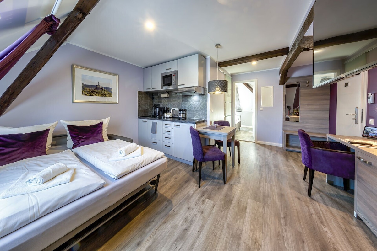 A modern bedroom with a large bed, a small kitchenette, and a dining area with purple chairs.