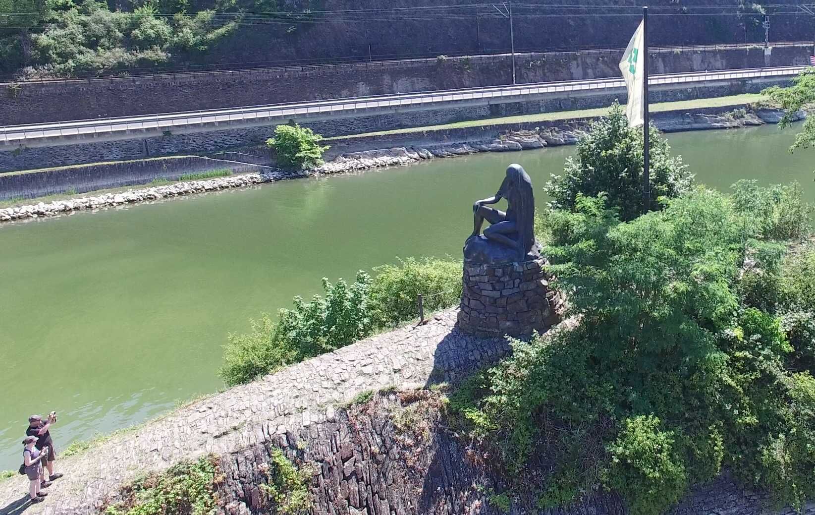 A statue on a stone platform overlooking a river, with a flagpole and greenery in the foreground.