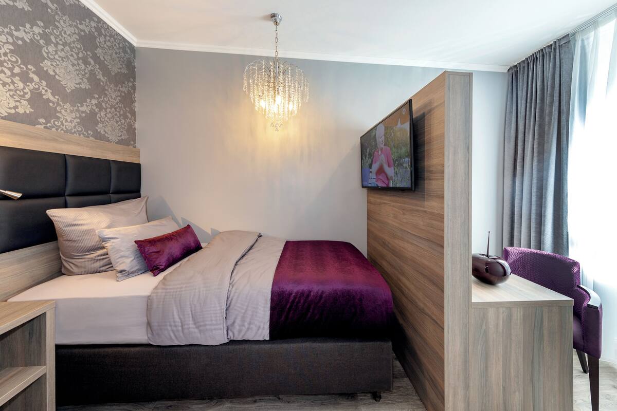 A modern hotel room with a bed, a desk, a chair, a chandelier, and a painting on the wall.