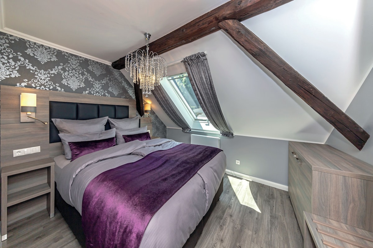 A bedroom with a sloped ceiling, a bed with a purple blanket, a wooden headboard, and a nightstand.