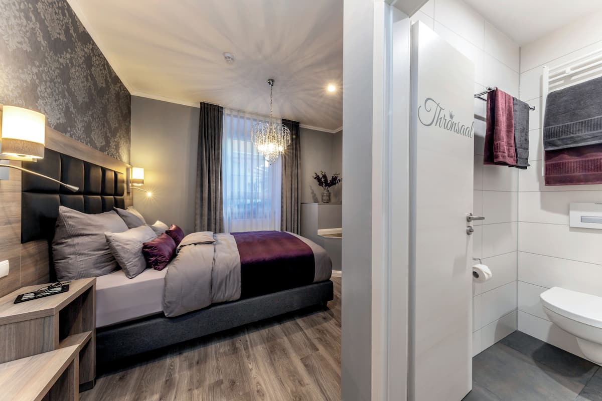 A modern bedroom with a bed, bedside tables, a chandelier, and a bathroom with a toilet and towel rack.