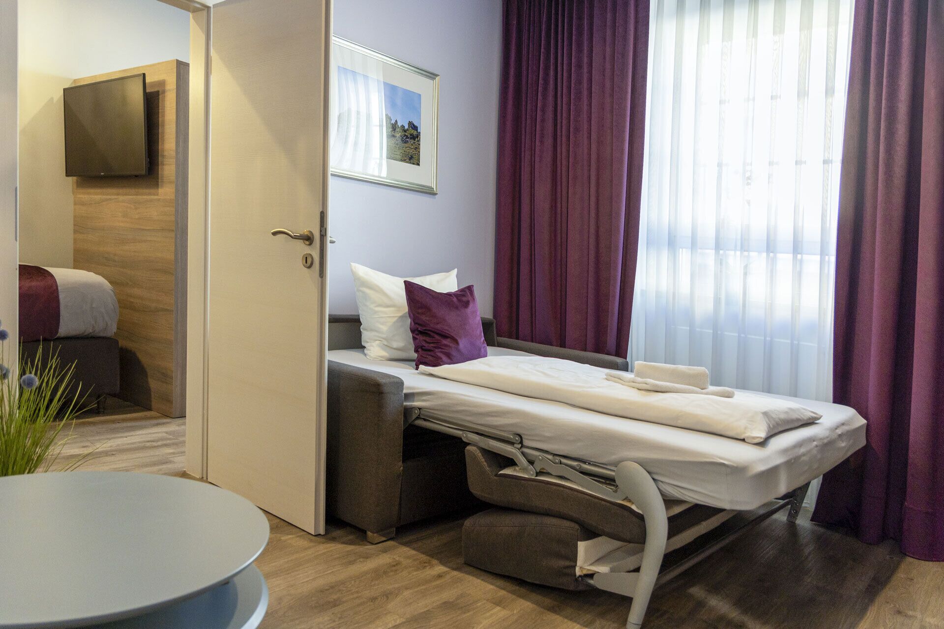 Una habitación de hotel moderna con una cama, una mesita de noche, una silla, un televisor montado en la pared y un ventanal con cortinas.