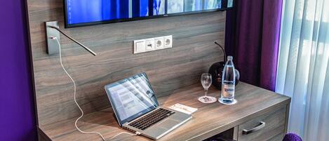 A modern hotel room with a wooden desk, a laptop, a glass of water, and a purple chair.