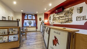 A reception area with a wooden counter, framed welcome sign, and a mural of a town scene. - Burgenheimat (Rhens)