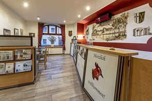 A reception area with a wooden counter, framed welcome sign, and a mural of a town scene. - Burgenheimat (Rhens)