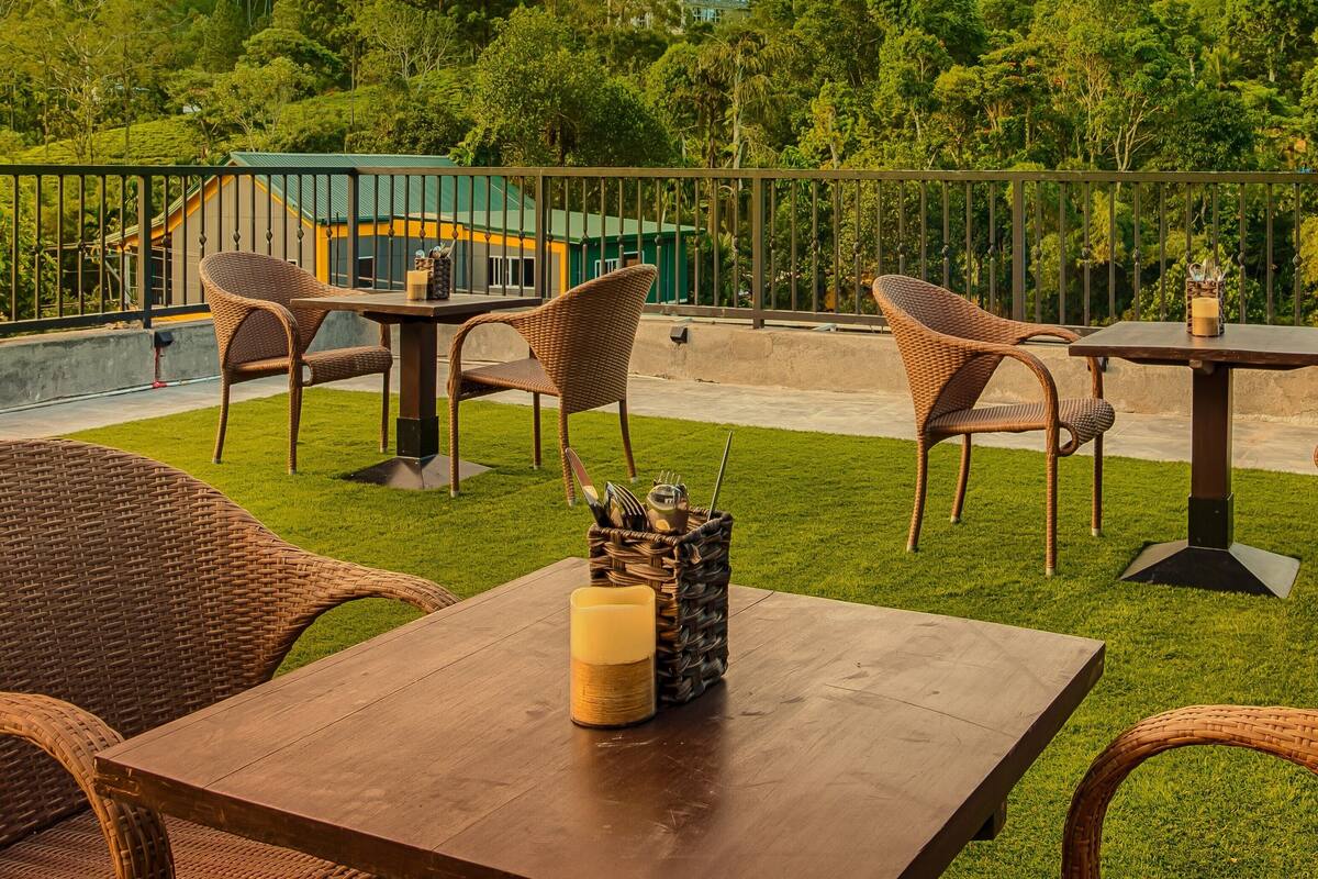 An outdoor patio with wicker furniture and a candle centerpiece, surrounded by lush greenery and a view of a building in the background.