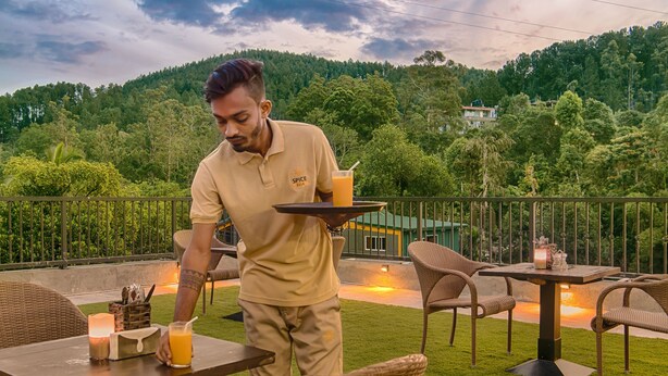 A person serving drinks at an outdoor table with wicker chairs, a grassy lawn, and a mountainous backdrop. - Spice Ella (Ella)