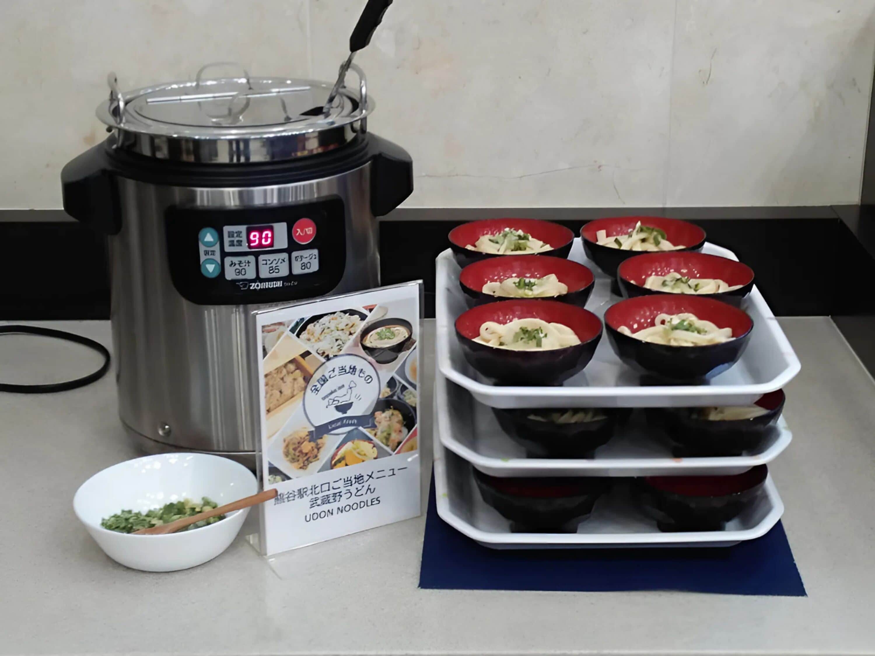 A food display with a pressure cooker, a bowl of green herbs, and a tray of red bowls with food.