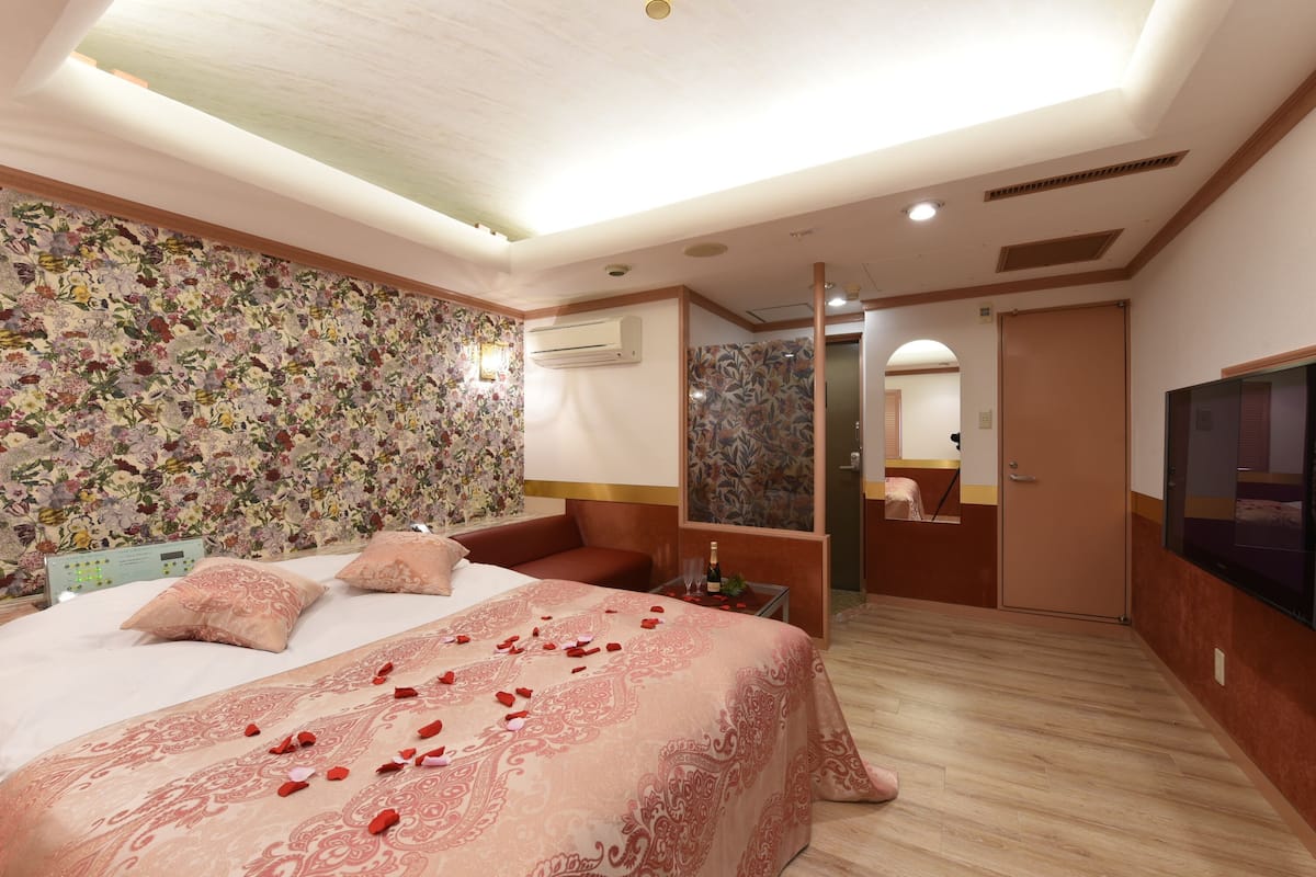 A hotel room with a floral wallpaper, a bed with rose petals, a TV, and a minibar.