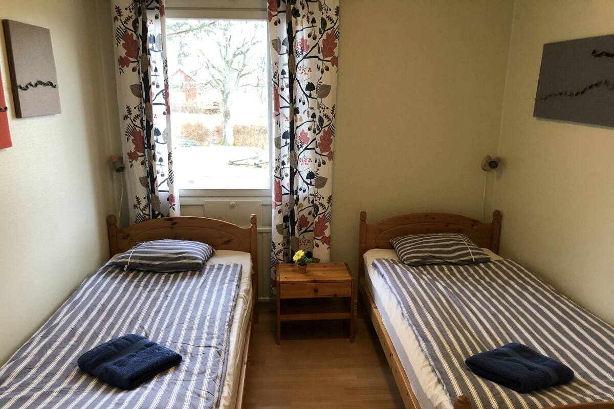 Ein Zimmer mit zwei Holzbett, jedes mit gestreifter Bettwäsche und einem blauen Handtuch. Zwischen den Betten befindet sich ein kleiner Nachttisch.