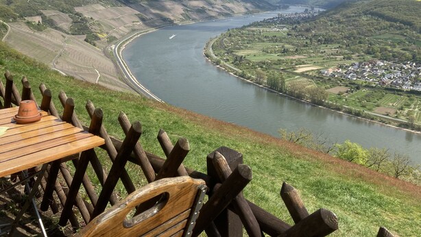 A scenic view of a river with a winding road alongside, a small town in the valley, and a wooden table with a chair in the foreground. - Haus Mittelrhein (Rhens)