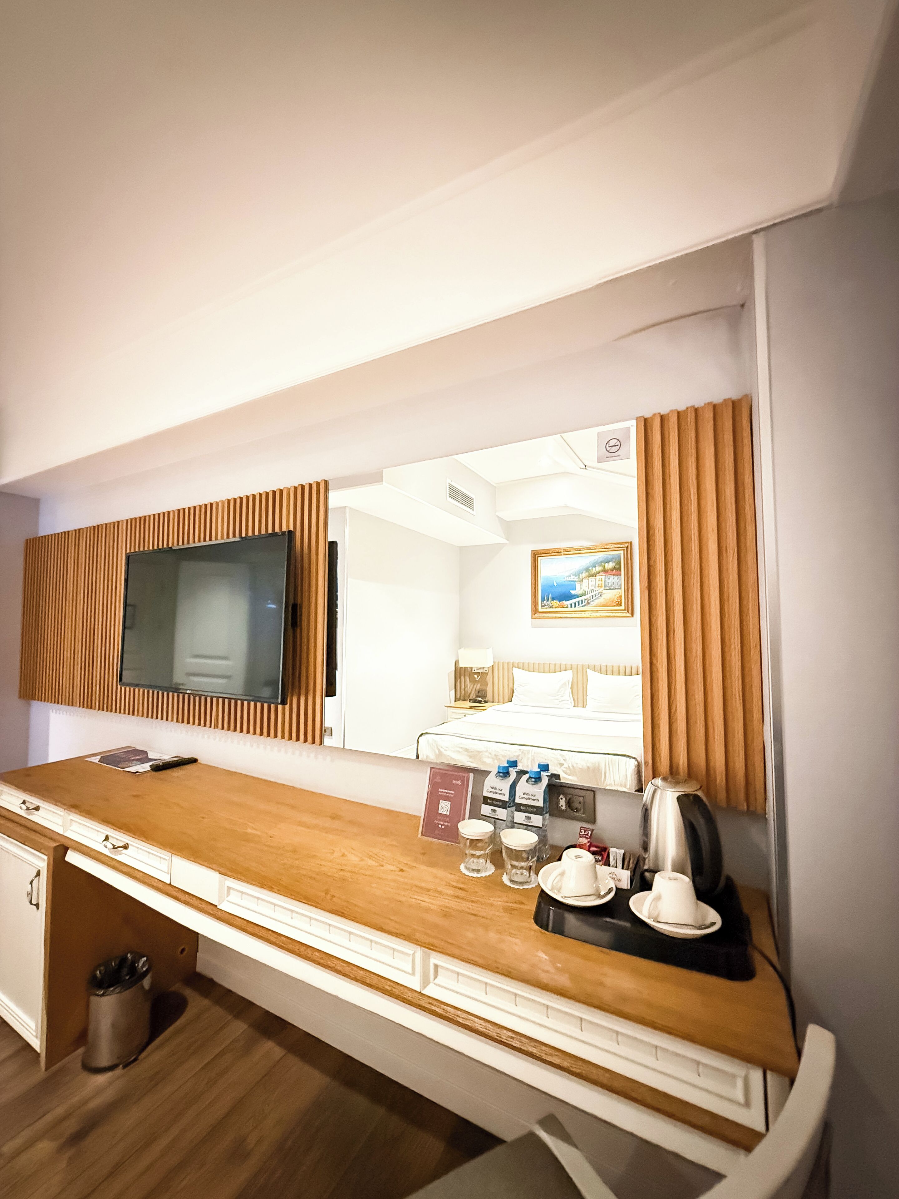 A hotel room with a wooden desk, a television, a bed, and a mirror reflecting the room.