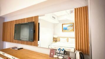 A hotel room with a wooden desk, a television, a bed, and a mirror reflecting the room.