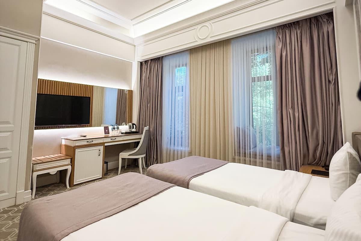 A hotel room with two beds, a desk, a chair, a television, and a large window with curtains.