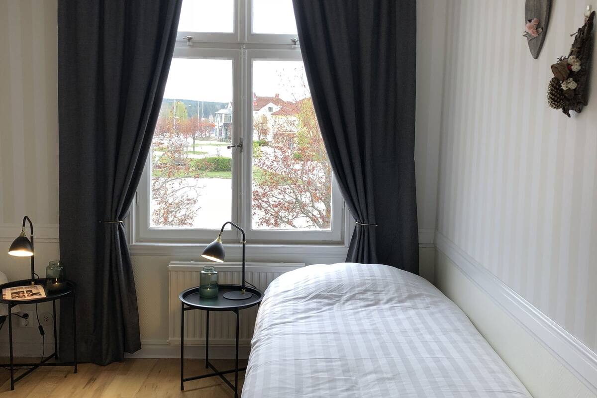 Un dormitorio con cama, ventana con cortinas, lámpara en una mesita de noche y vistas al exterior.