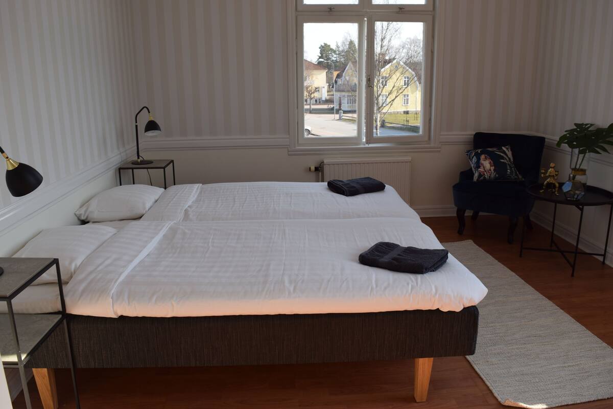 Un dormitorio con dos camas, una ventana que ofrece vistas al exterior, una mesita con una lámpara y una silla con un cojín decorativo.