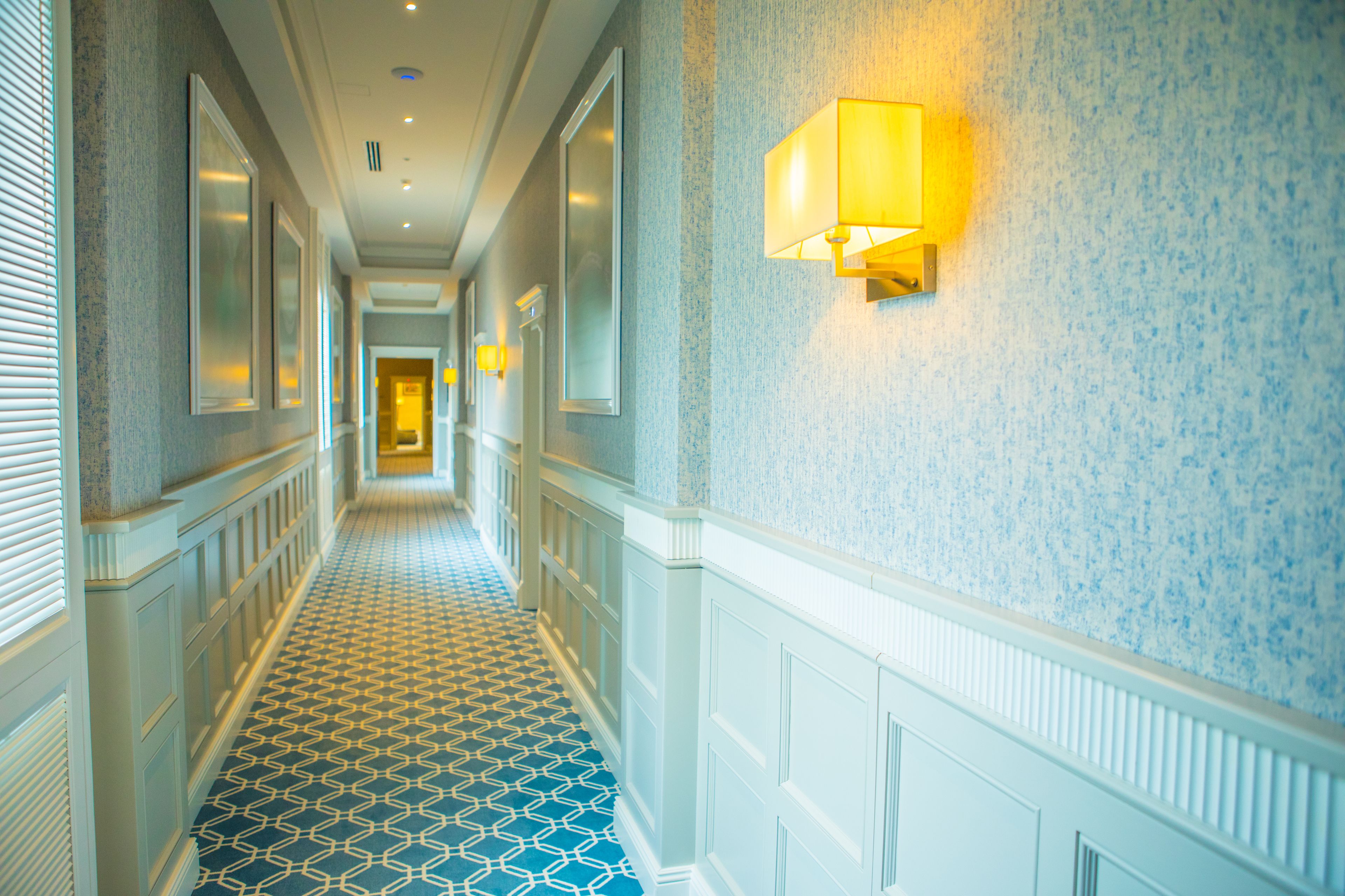 A hotel hallway with patterned flooring, wall-mounted lamps, and framed mirrors.