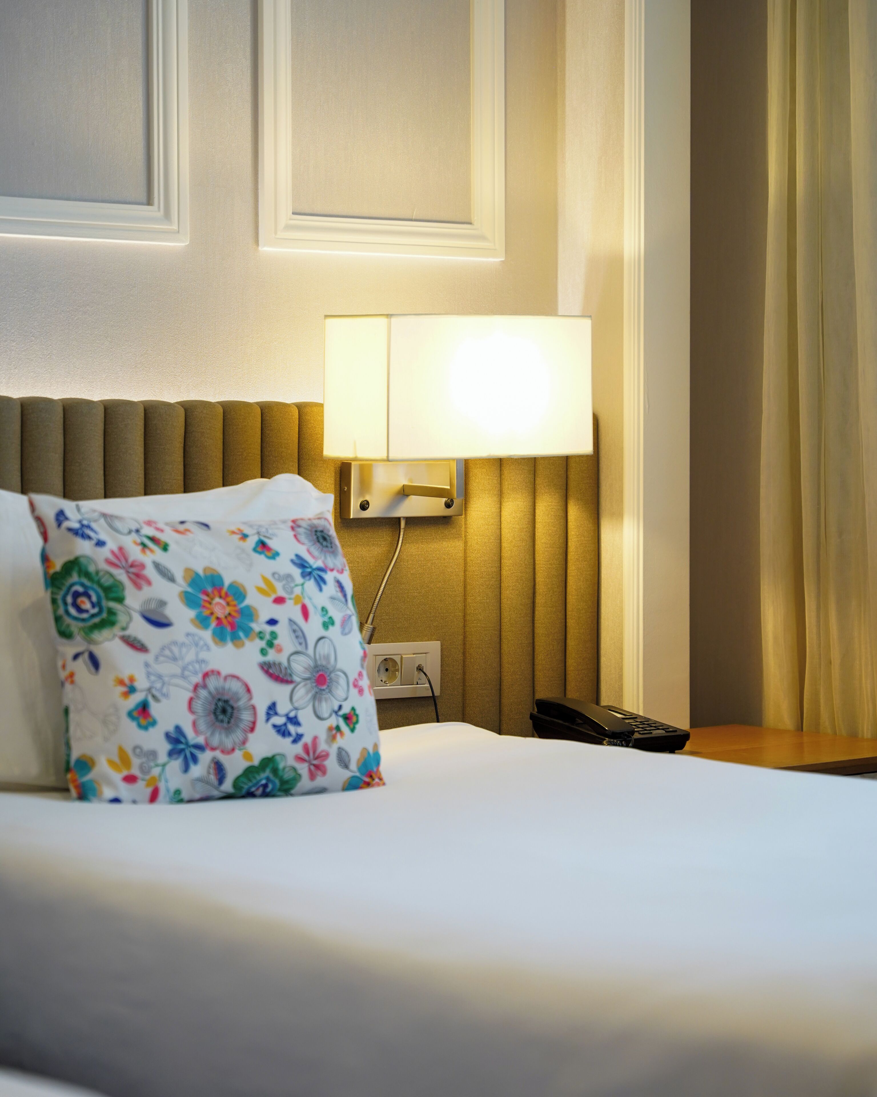 Una camera d'albergo con un letto, una lampada da comodino, un telefono e un cuscino floreale.