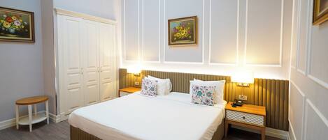 A hotel room with a large bed, bedside tables, a nightstand, a small stool, and two framed floral paintings on the wall.