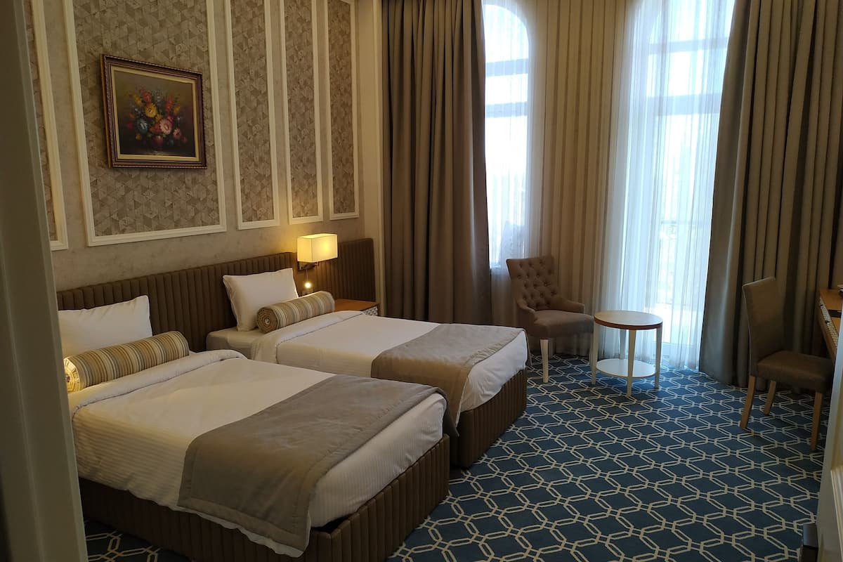 A hotel room with two beds, a small table, a chair, and a window with curtains.
