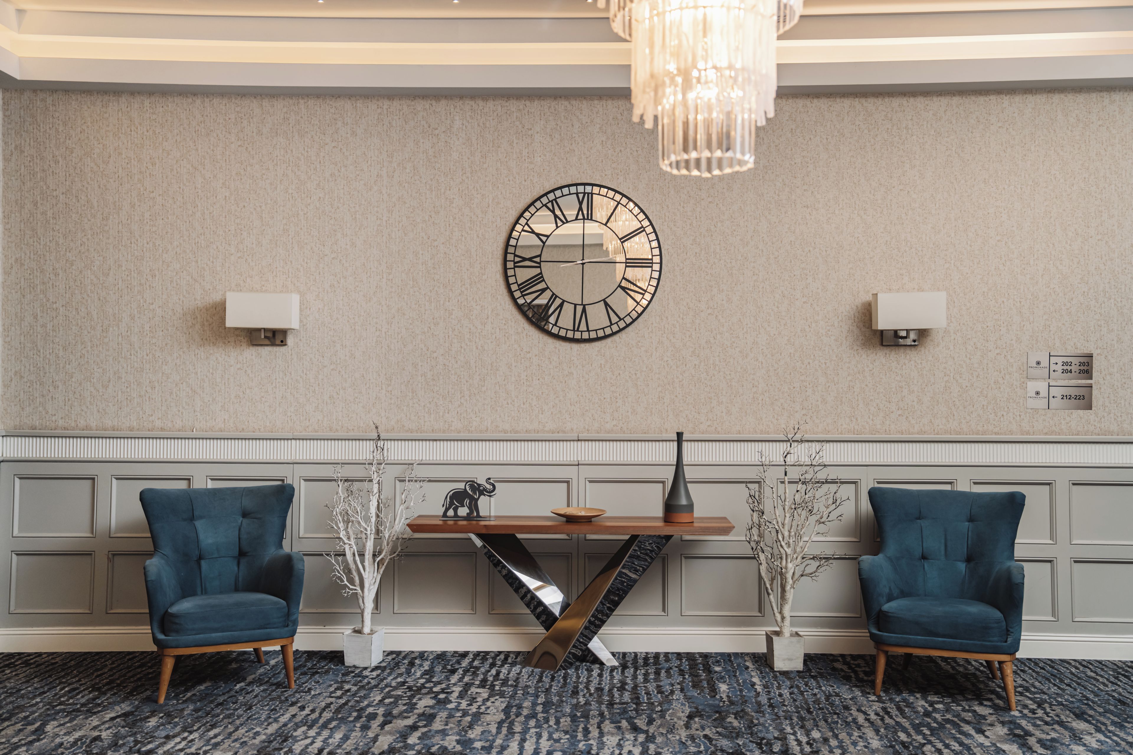 A hotel lobby with a large wall clock, two armchairs, a wooden table, and a chandelier.