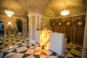 A lobby with a marble reception desk, chandeliers, a clock, and a check-in counter.