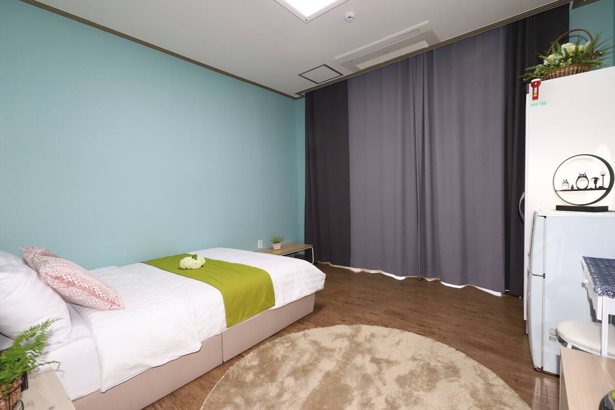 A bedroom with a bed, a desk, a chair, a small table, a plant, and a window with curtains.