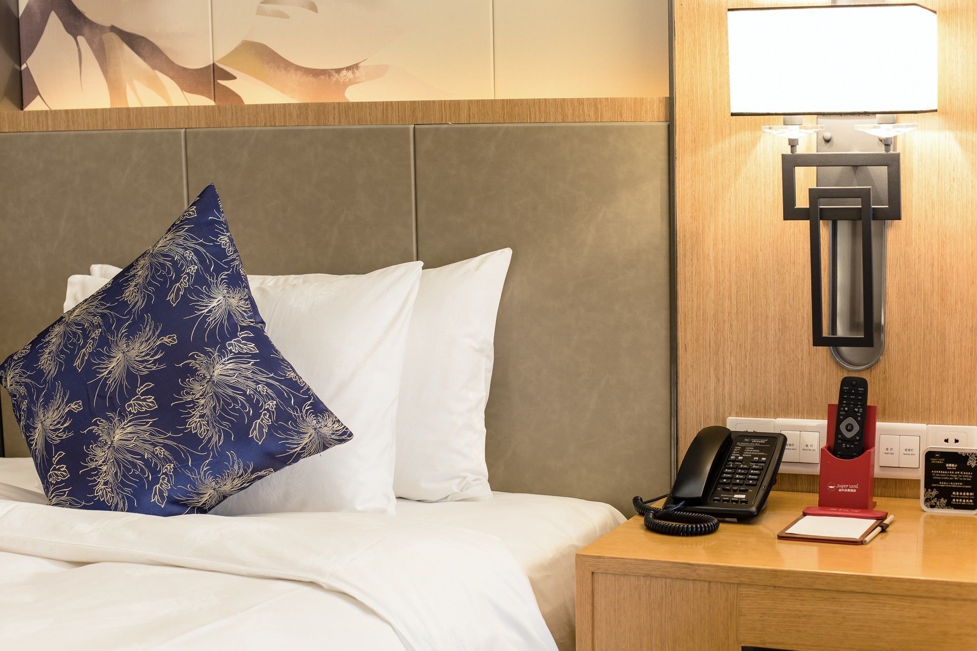 A hotel room with a bed, white pillows, a blue floral pillow, a bedside table with a telephone, a red telephone, and a wall-mounted light.