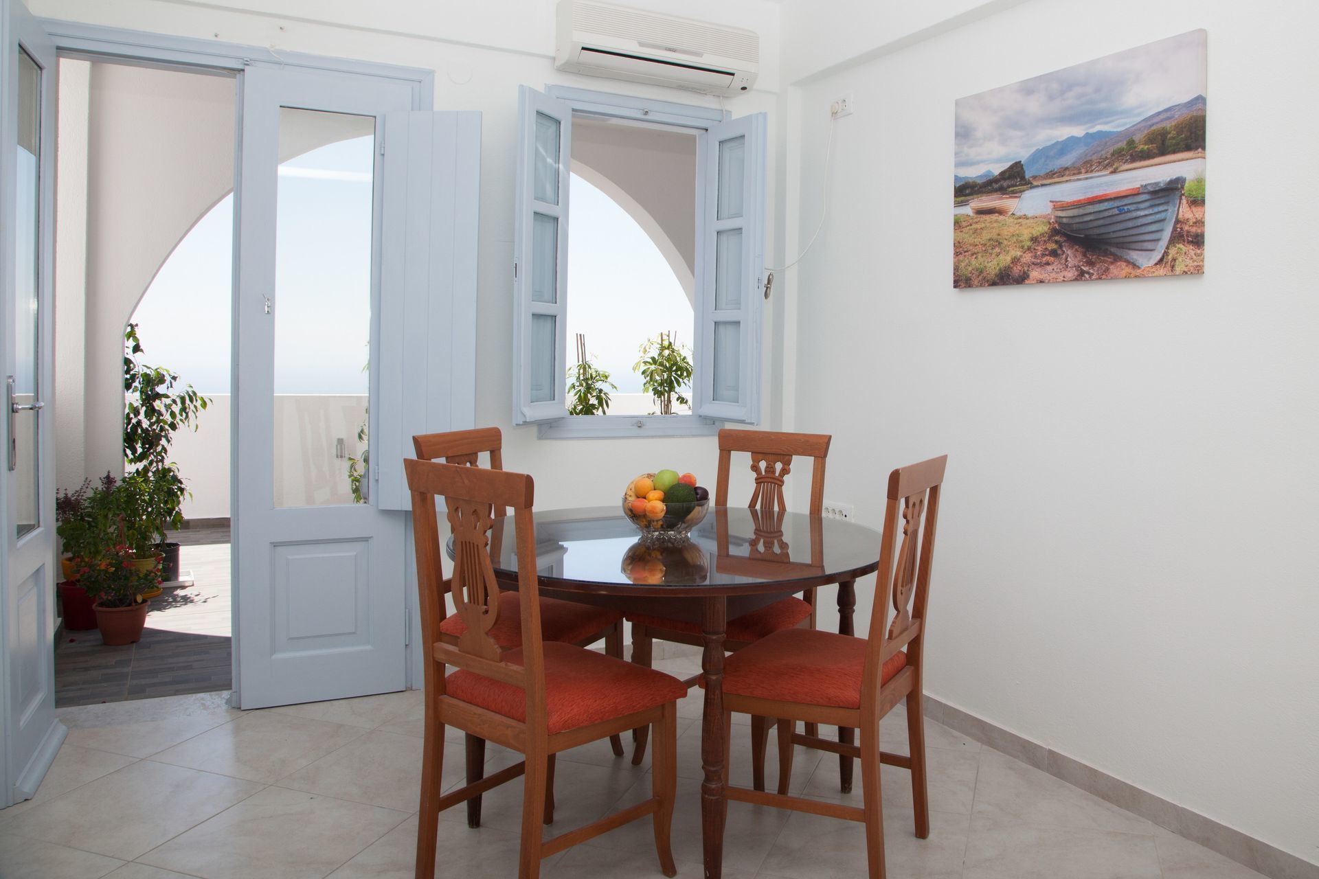A dining area with a round table, four wooden chairs with red cushions, and a bowl of fruit. There are open doors leading to a balcony with potted plants.