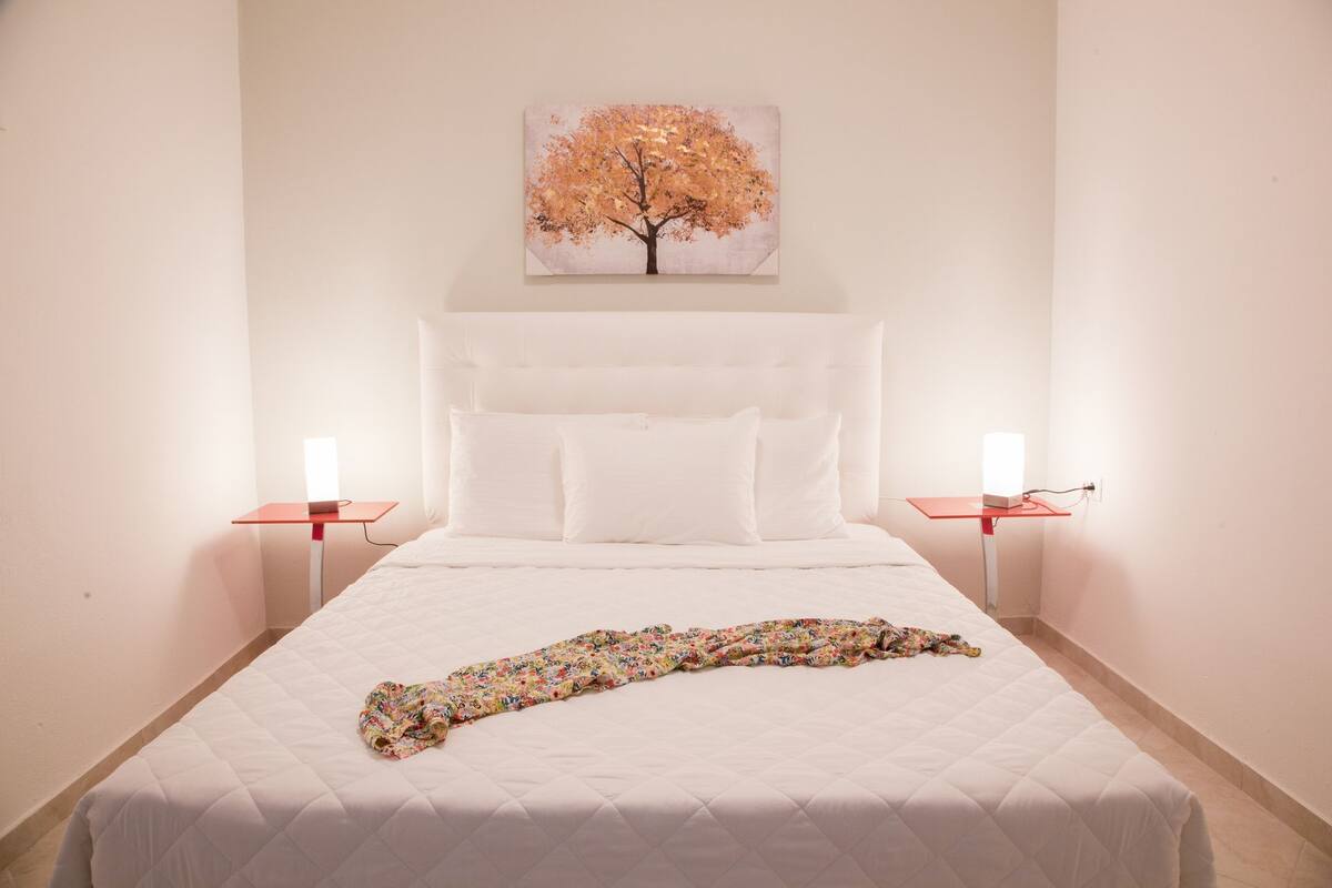 A bedroom with a large bed, white bedding, a floral blanket, two bedside tables with lamps, and a painting of a tree on the wall.