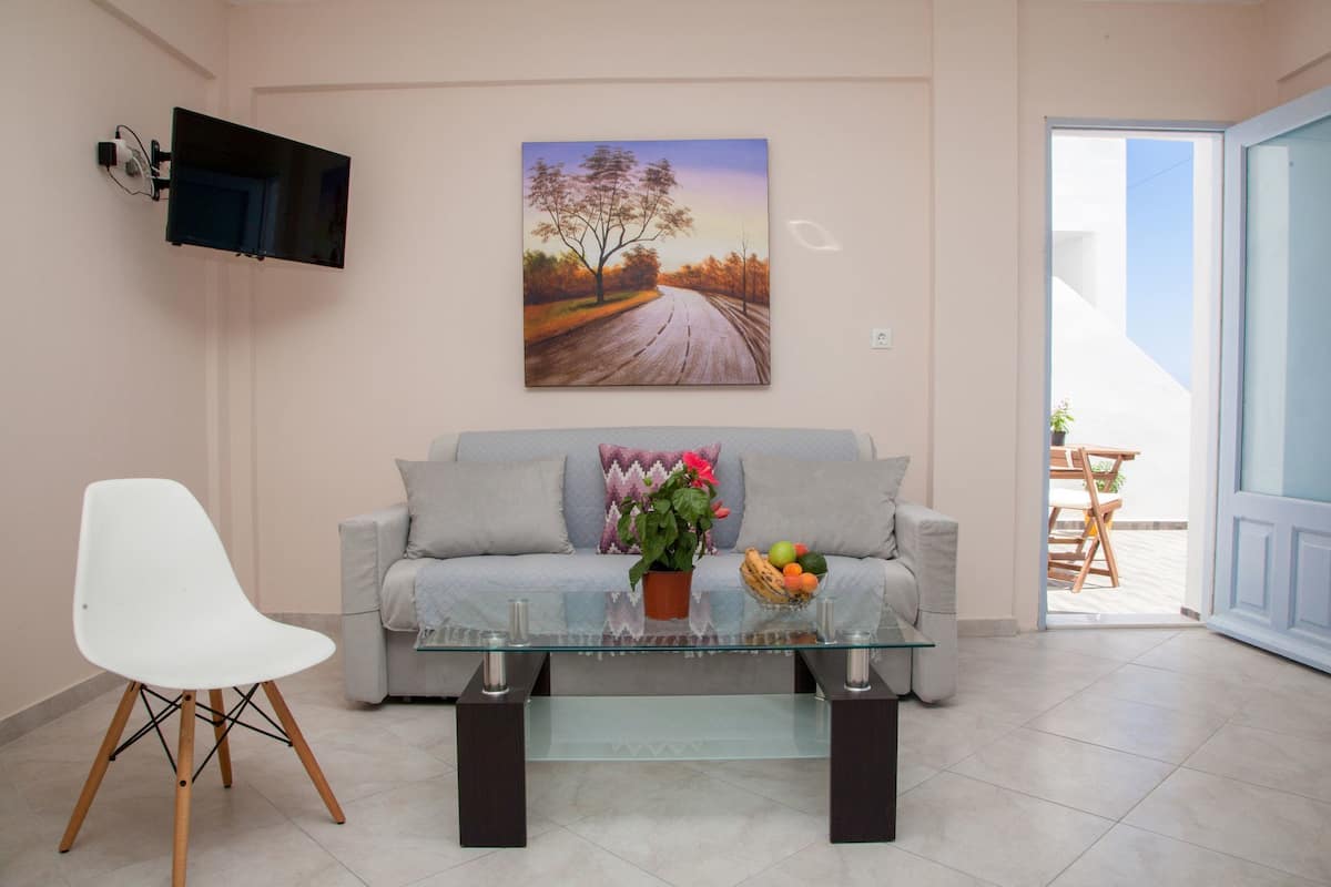 A living room with a sofa, a glass coffee table, a white chair, a TV mounted on the wall, and a painting above the sofa.