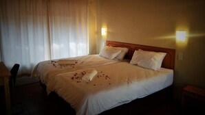 A double bed with white linens, two pillows, and a rolled towel on the bed. There are two bedside lamps and a window with curtains. - Ananda Lodge (Rustenburg)