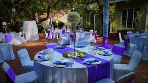An outdoor wedding reception area with round tables covered in white tablecloths and purple sashes, surrounded by white chairs. - Ananda Lodge (Rustenburg)