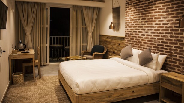 Habitación de hotel con una cama de madera, un escritorio con silla, una mesita pequeña y una pared de ladrillo.
