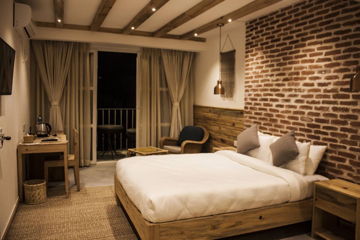 Habitación de hotel con una cama de madera, un escritorio con silla, una mesita pequeña y una pared de ladrillo.