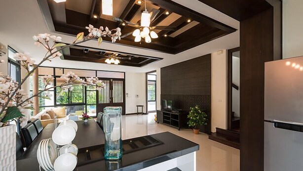A modern kitchen with a black countertop, white plates, and a clear vase. There is a dining area with a table and chairs, and a large window with a view of greenery. - Pool Villa Pattaya by Passionata (Pattaya)