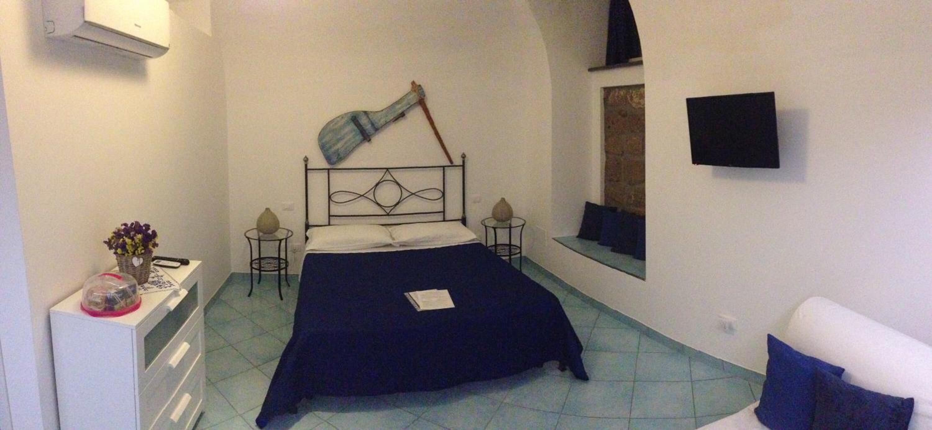 Una habitación pequeña y estrecha con una cama, una mesita con un cesto de flores, una silla y una ventana.