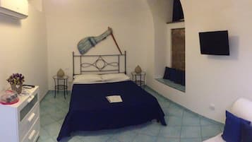 Una habitación pequeña y estrecha con una cama, una mesita con un cesto de flores, una silla y una ventana.