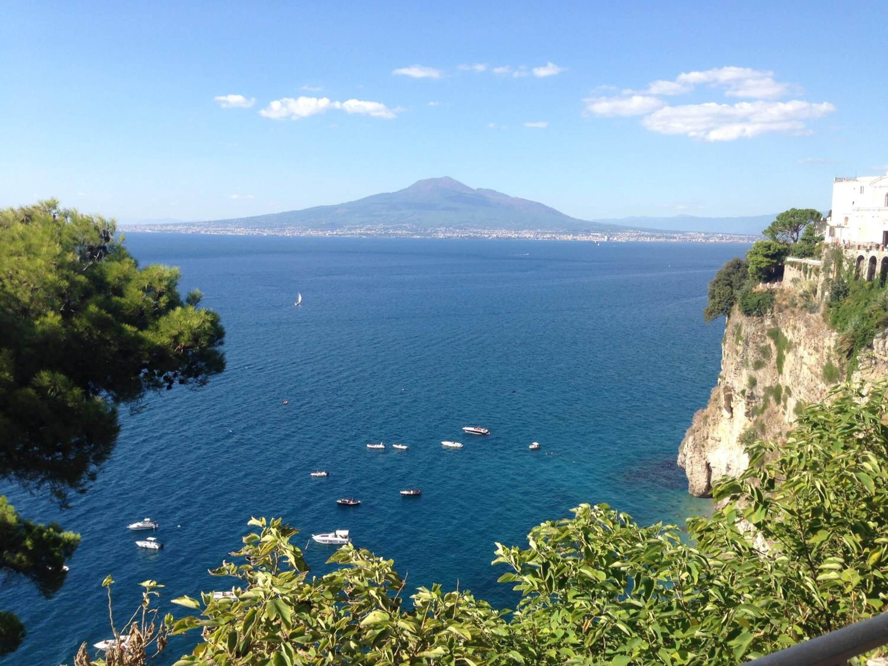 Vista costera con barcos en el agua, una montaña al fondo y un edificio en el acantilado.