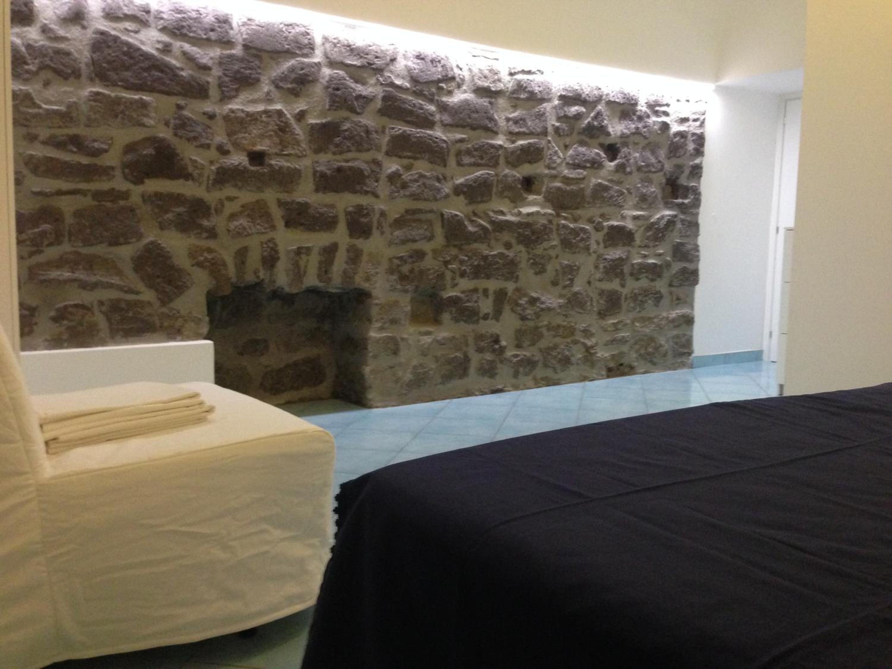 A room with a stone wall, a bed, a chair, and a small fireplace.