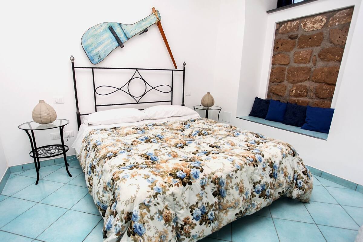 A bedroom with a bed, a guitar, a window with a brick wall, and a small table with a vase.