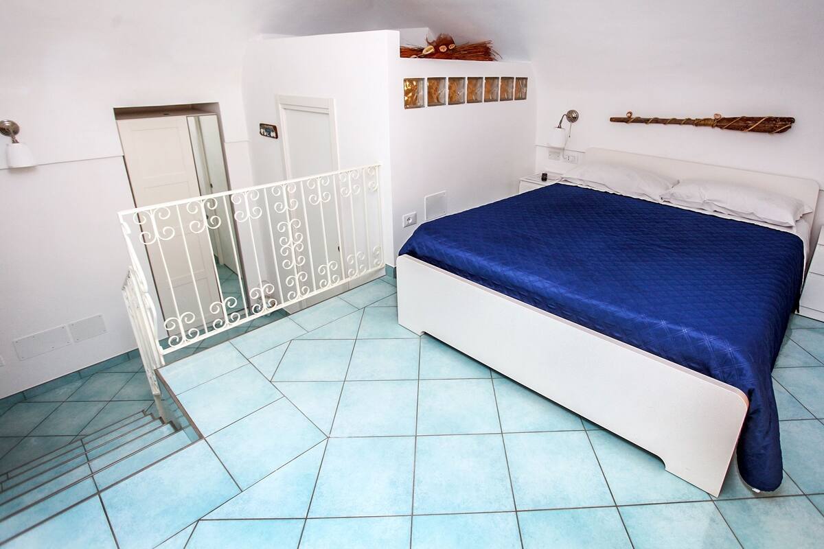 A bedroom with a bed, white walls, a white door, a white railing, and a blue floor.