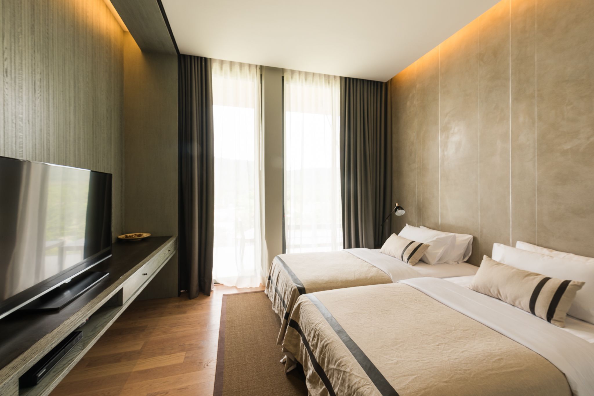 Quarto de hotel com duas camas, televisão de tela plana, piso de madeira e janelas amplas com cortinas.