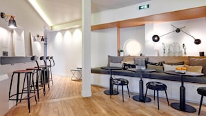 A modern interior with a wooden floor, bar stools, a seating area with a round table, and a wall-mounted light fixture. - Tinah Paris - Reaumur Montorgueil (Paris)