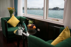 Quarto com sofá verde, uma pequena mesa com uma fruteira e copos, e vista para um corpo d'água através de grandes janelas.