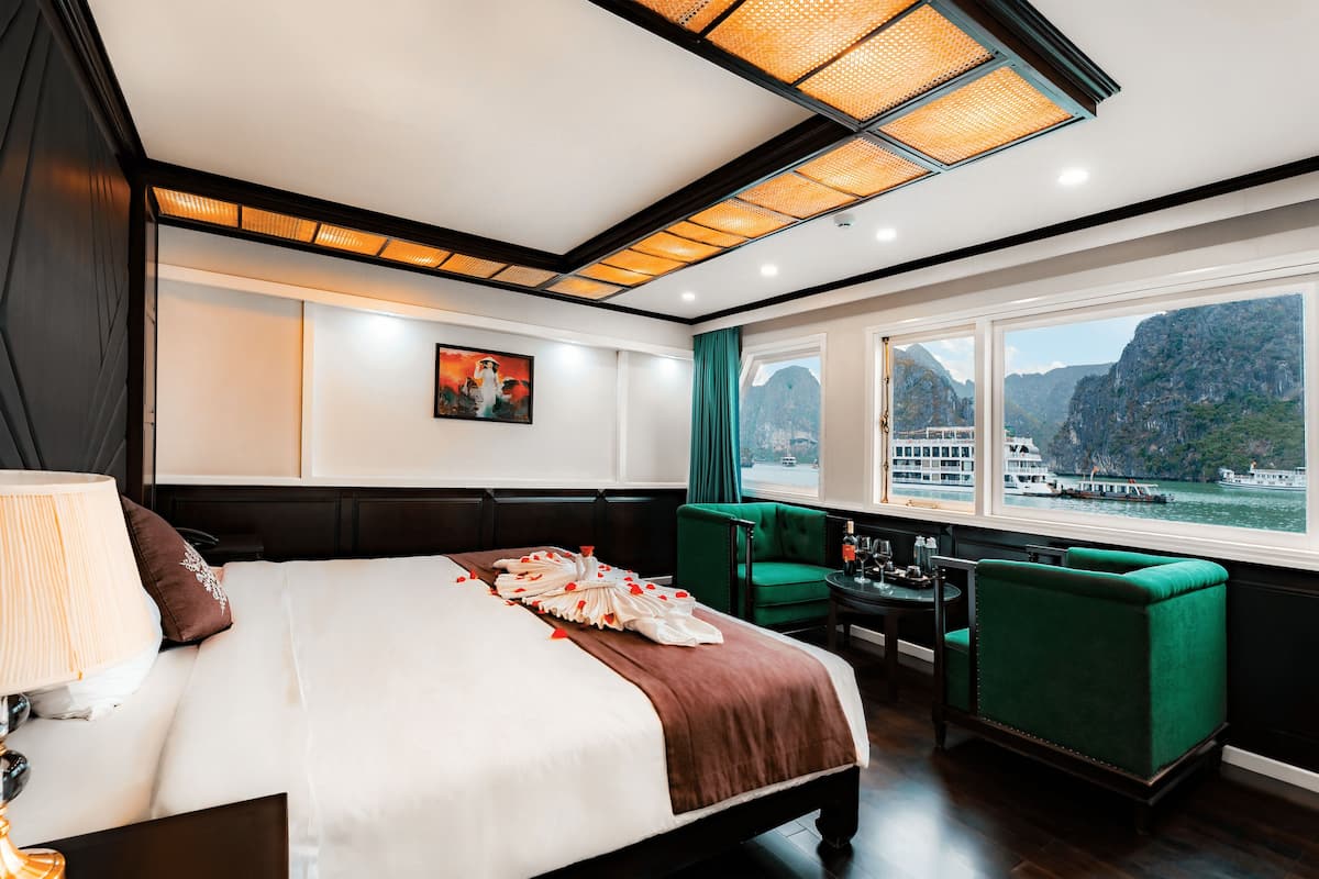 A hotel room with a large bed, a view of a mountainous landscape, a green armchair, and a small table with a bottle and glasses.