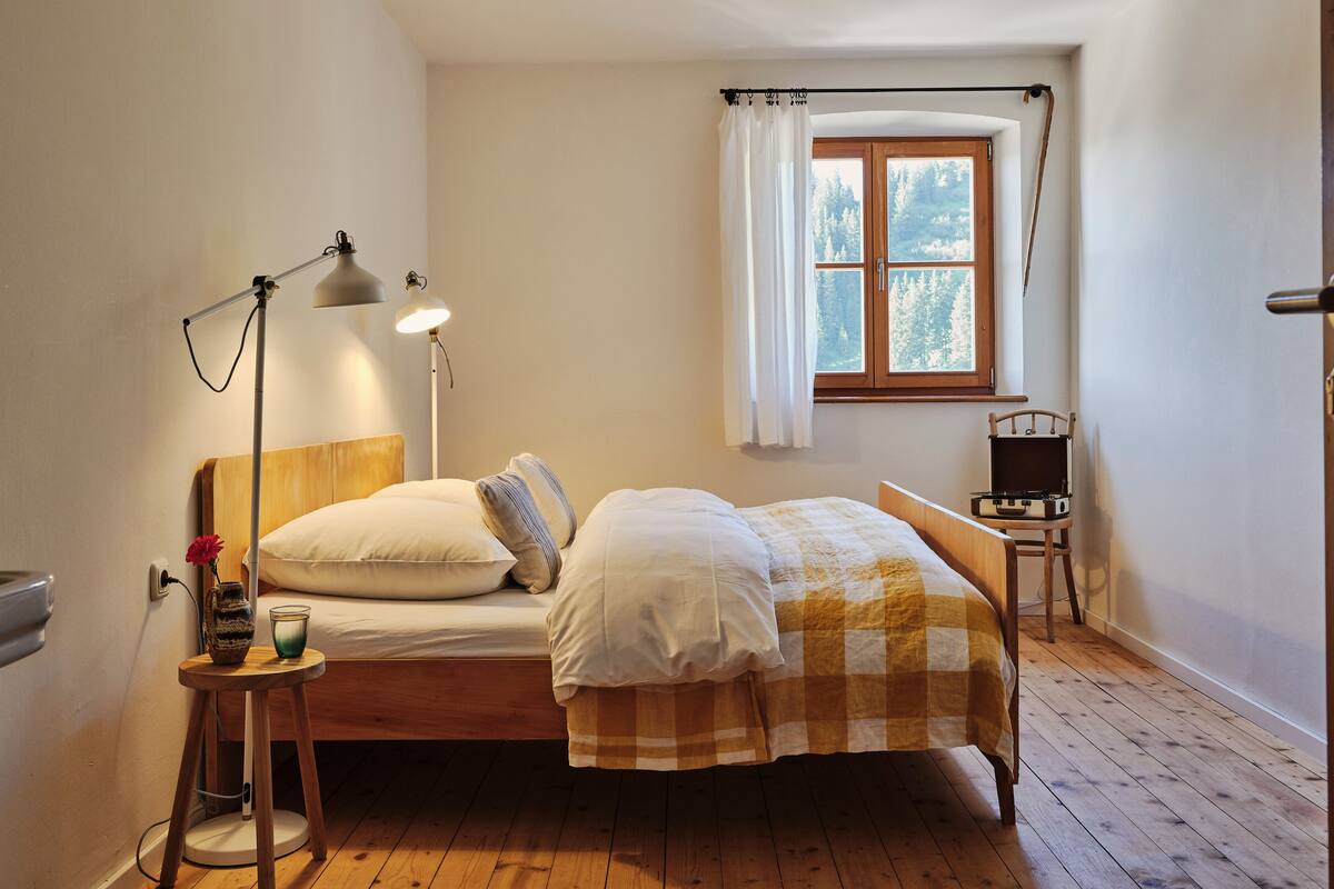 A bedroom with a wooden bed, a bedside table with a lamp, a window with curtains, and a view of trees outside.